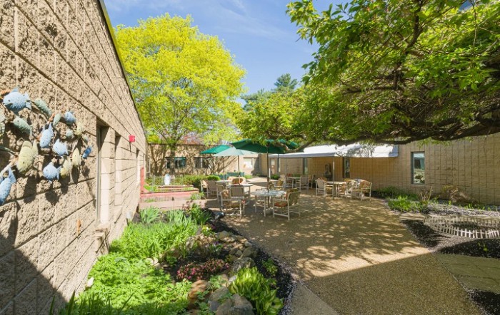 A tranquil courtyard featuring outdoor seating with tables and chairs, surrounded by lush greenery on a sunny day.