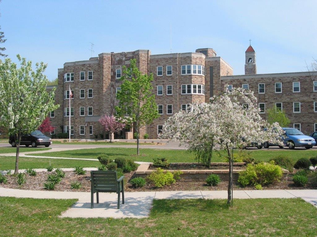 A university campus featuring a large stone building surrounded by trees and a bench in the foreground.
