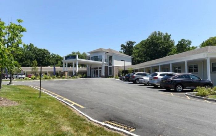 A modern building with a parking lot and several parked cars surrounded by greenery on a sunny day.