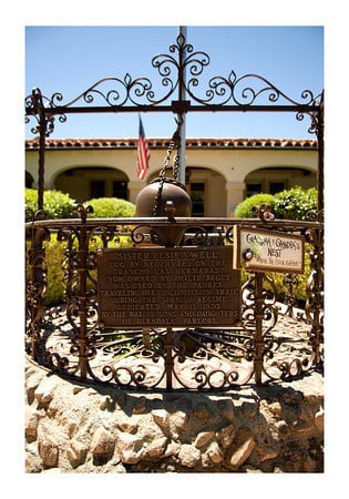 A historic site marked with a plaque, surrounded by an ornate iron gate, with a building and garden in the background.