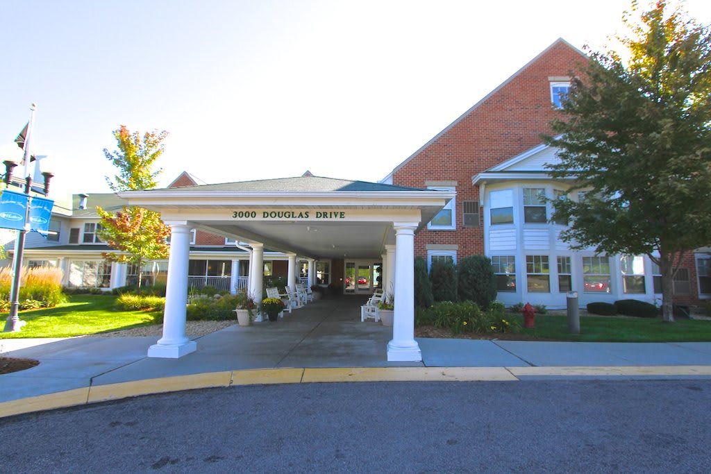 A building entrance with columns and a driveway, surrounded by landscaping and bathed in sunlight.