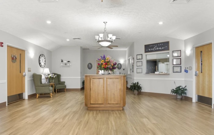 A modern reception area featuring wooden flooring, a central desk with a floral arrangement, and a waiting area with seating.