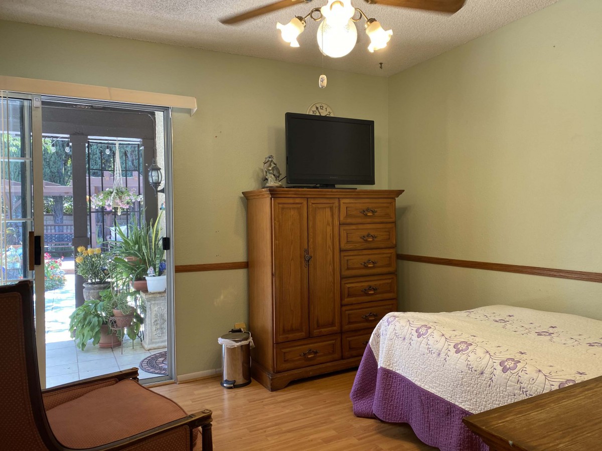 A cozy bedroom featuring a wooden dresser with a TV on top, a bed with a quilt, and sliding glass doors leading to a space with indoor plants.
