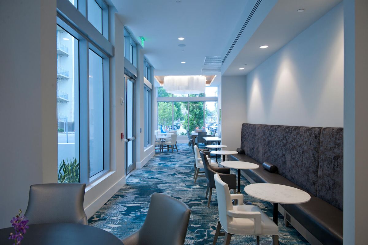 A modern cafe interior with seating areas, featuring multiple tables and chairs, illuminated by natural light through large windows.
