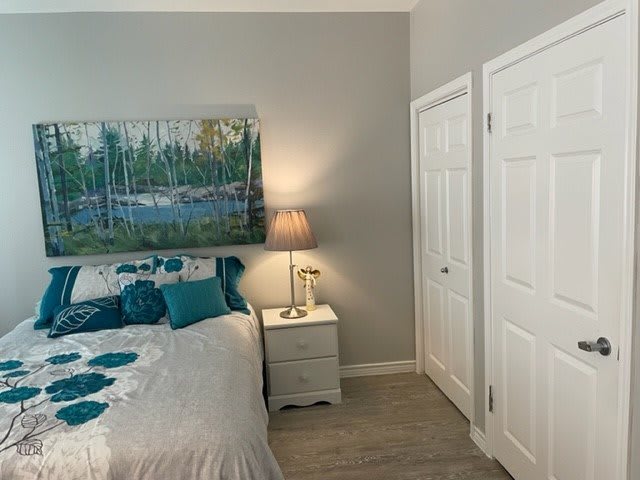 A cozy bedroom featuring a bed with teal pillows, a bedside lamp, and a landscape painting on the wall.