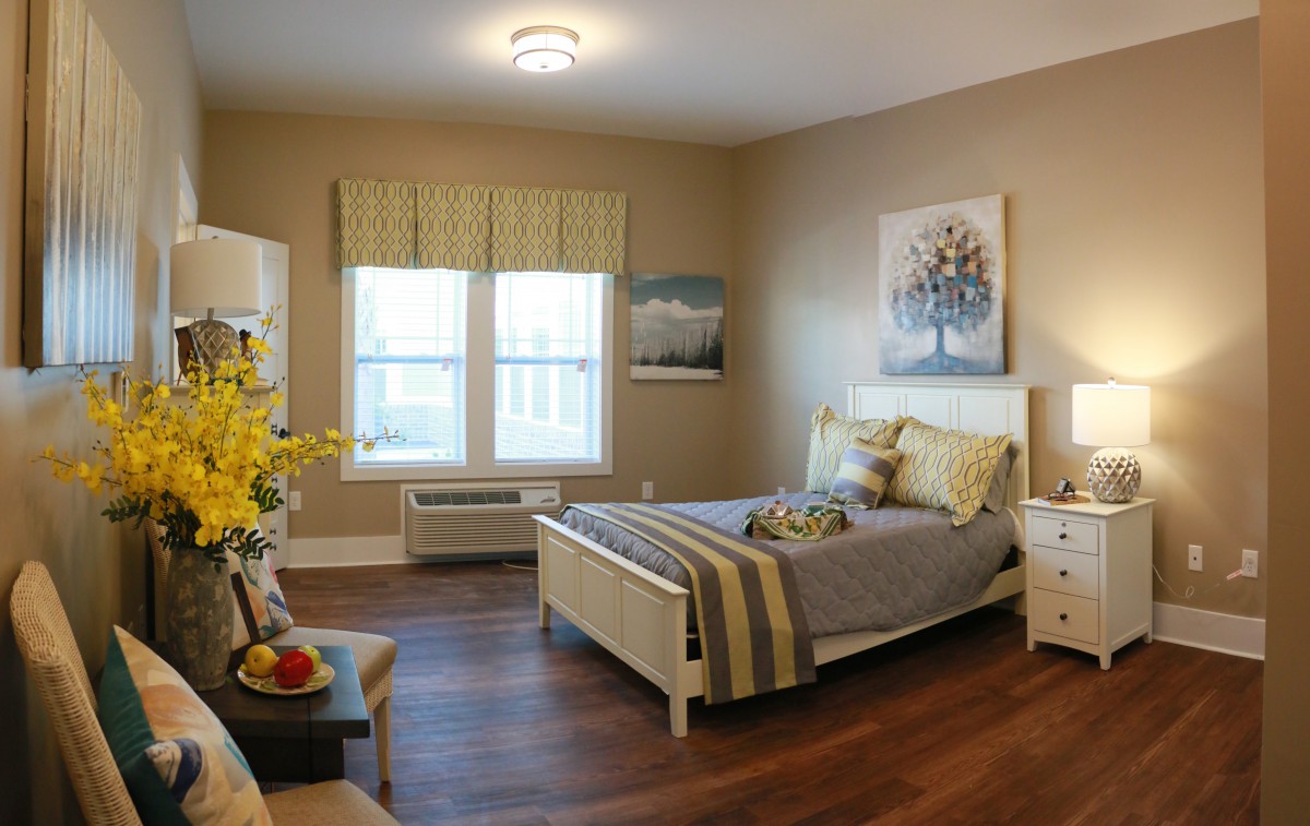 A cozy and well-decorated bedroom with natural light streaming in through two windows, featuring a bed with decorative pillows, wooden flooring, and tasteful wall art.