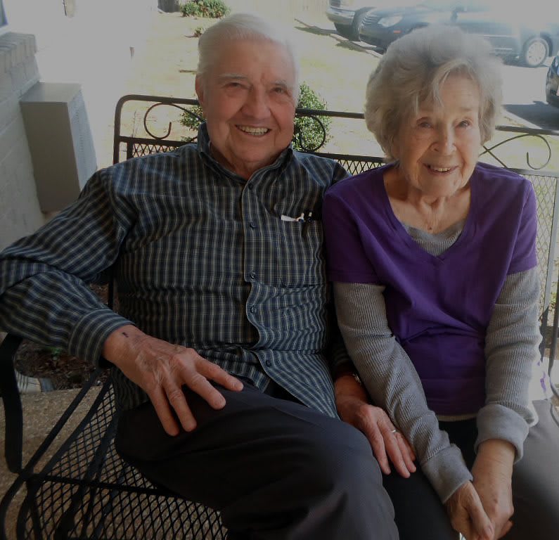 An elderly couple sitting together on a bench, smiling outdoors.