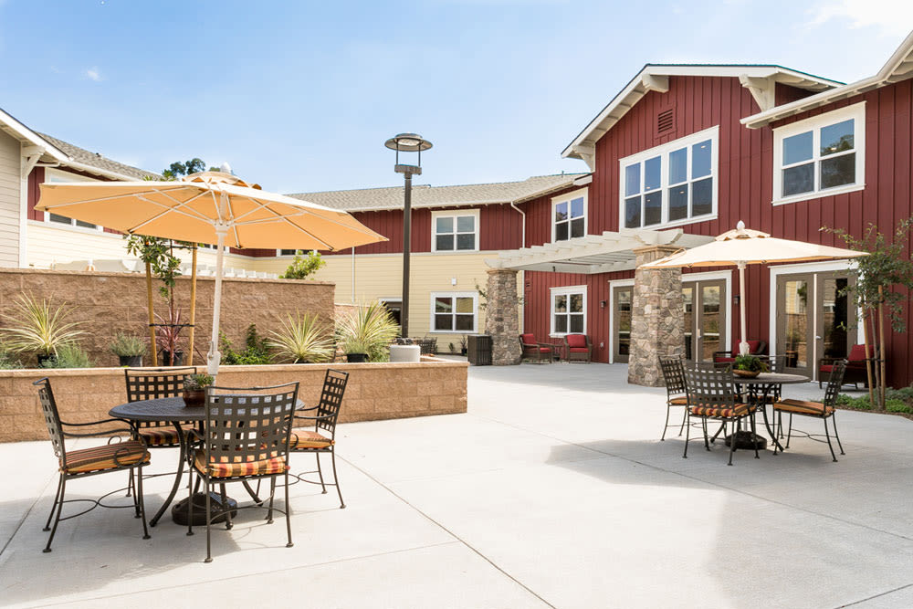 A sunny outdoor patio area with tables, chairs, and umbrellas in front of a red building.