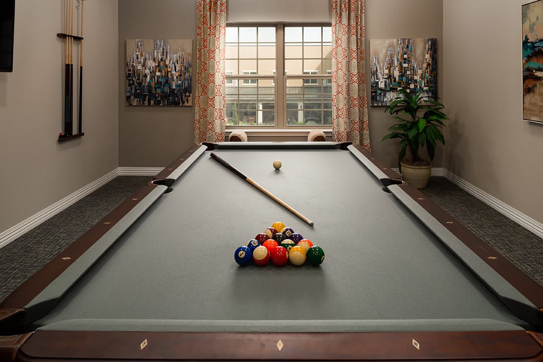 A well-decorated billiard room featuring a pool table with neatly arranged balls and a cue stick, set against a backdrop of large windows and artwork.