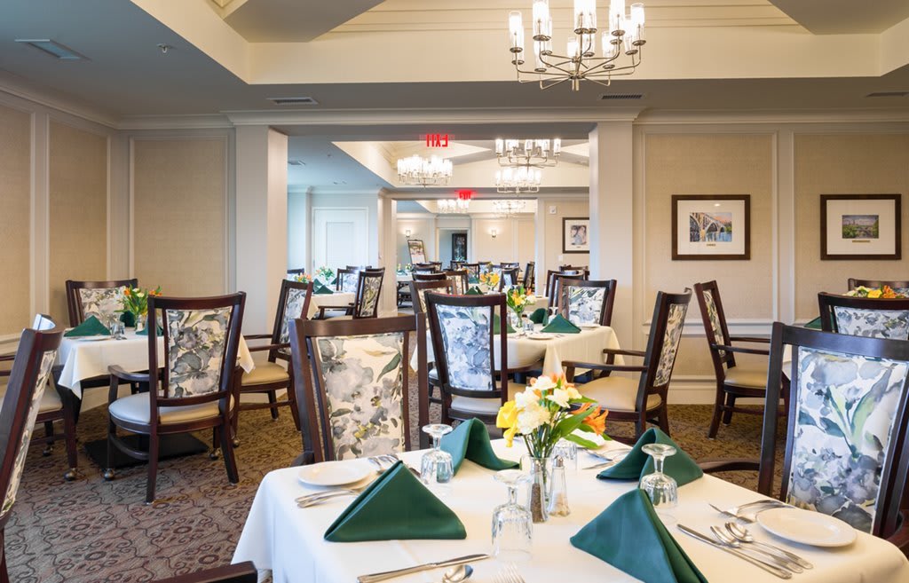 An elegant dining room with neatly set tables, featuring green napkins and floral centerpieces.
