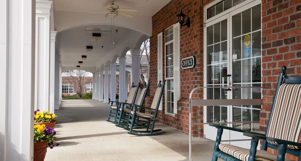 A row of rocking chairs on a covered porch of a brick building.