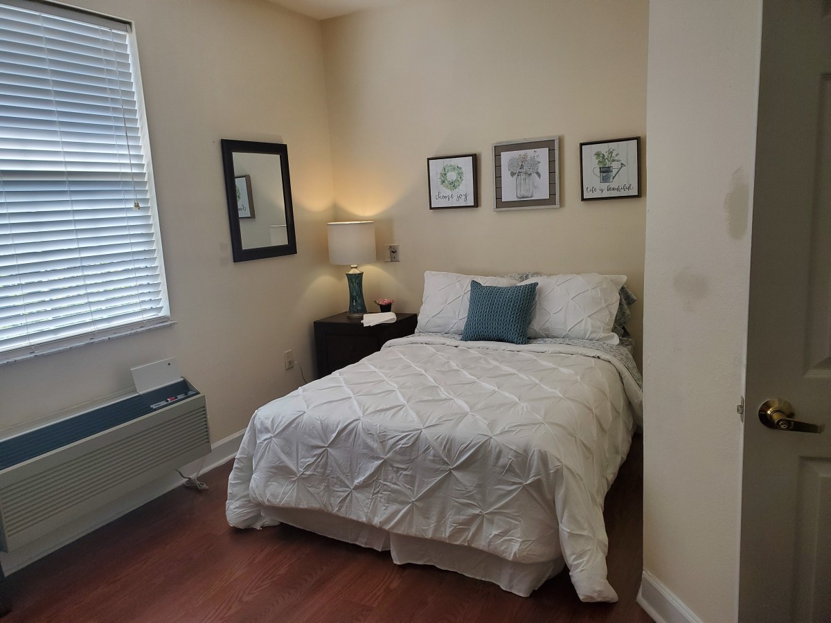 A cozy bedroom with a neatly made bed, decorative pillows, framed artwork on the wall, and a lamp on the bedside table.