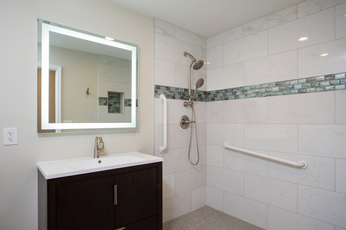 A modern bathroom featuring a vanity with an illuminated mirror, a tiled shower area with a handheld showerhead, and grab bars for safety.