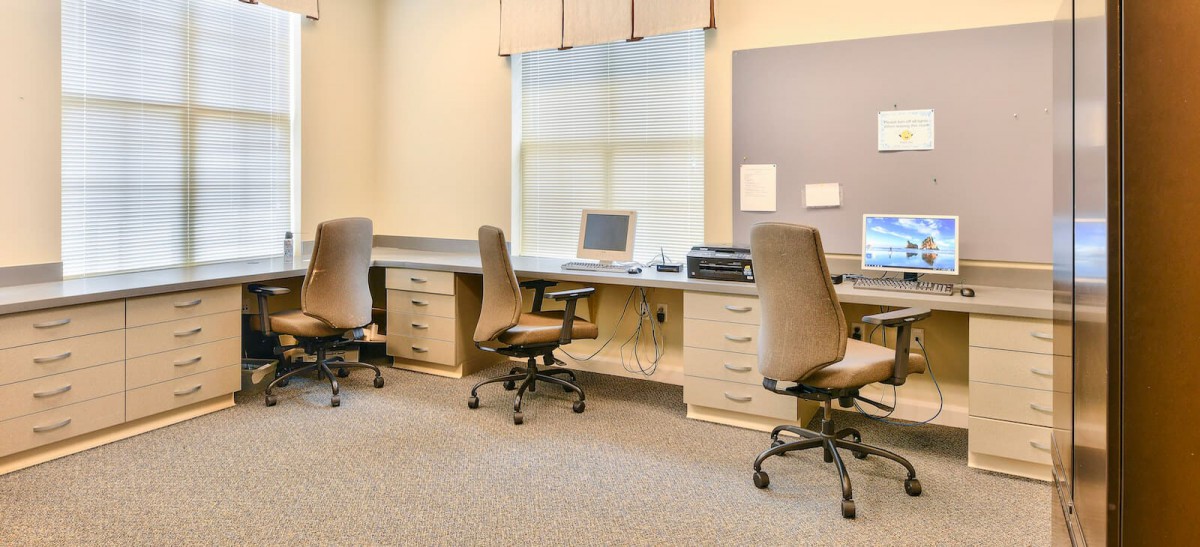 A modern office workspace with multiple desks and chairs, equipped with computers and a window view.