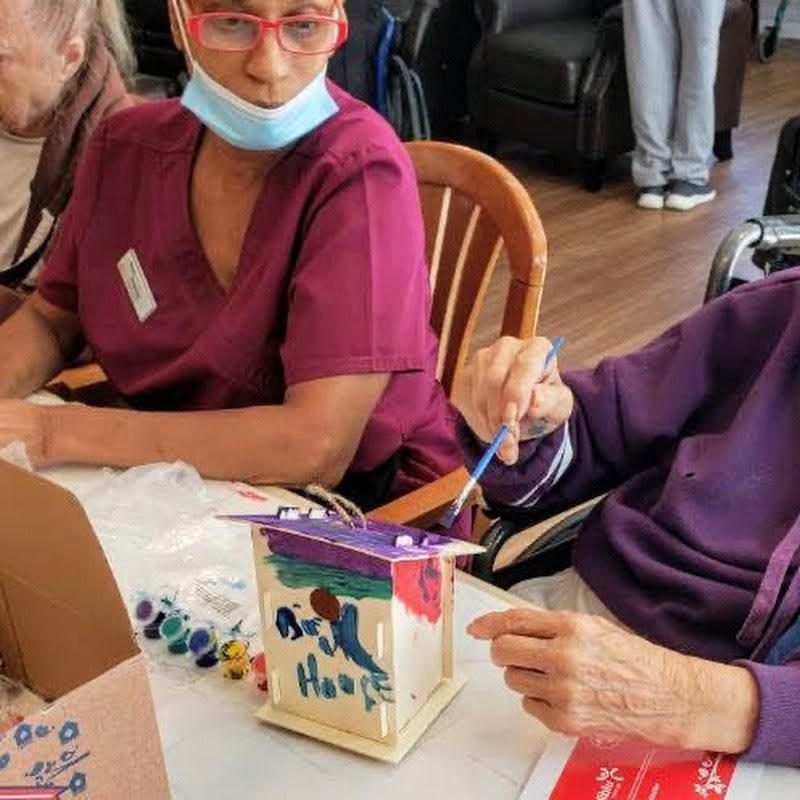 Elderly person painting a birdhouse with assistance from nursing staff.