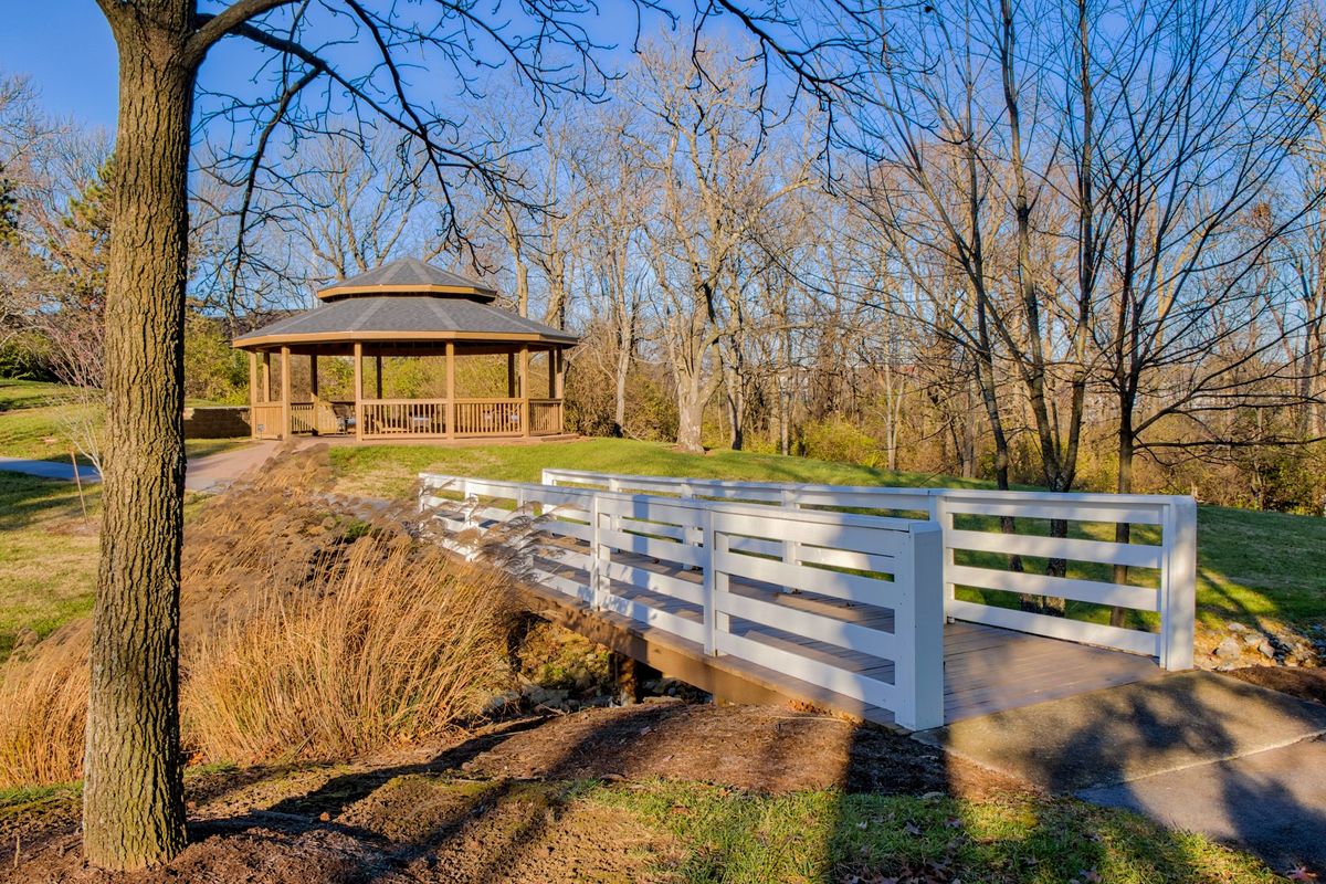 A serene park scene with a white bridge leading to a wooden gazebo surrounded by trees.