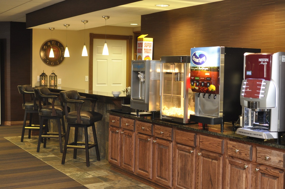 A cozy concession area with a popcorn machine and soda dispenser, featuring wooden cabinets and bar stools.