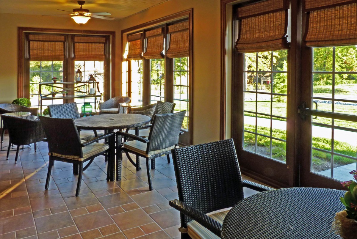A cozy sunroom dining area with multiple tables and chairs, featuring large windows and blinds.