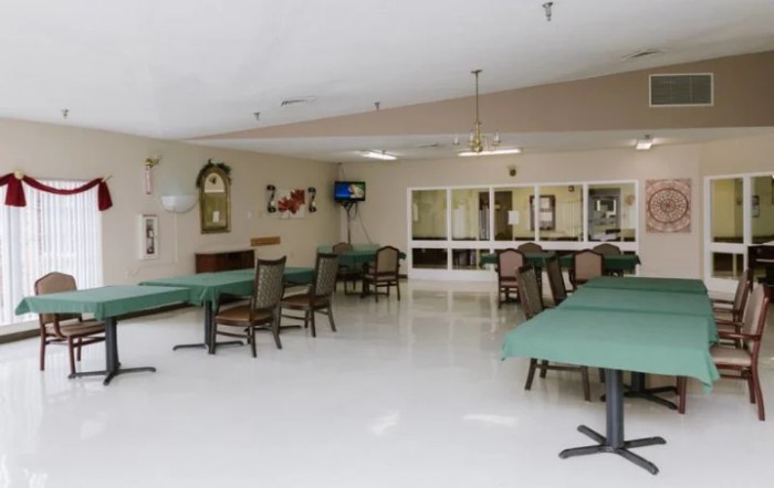 An empty dining area with green tablecloth-covered tables and chairs, featuring a TV and mirrors on the wall.