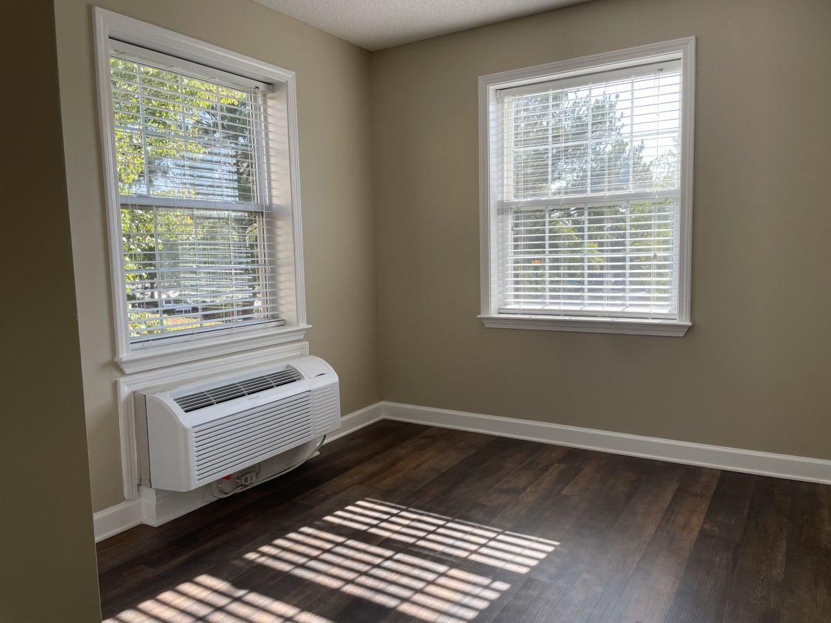 A sunlit empty room with hardwood flooring and two windows, featuring an air conditioner unit.