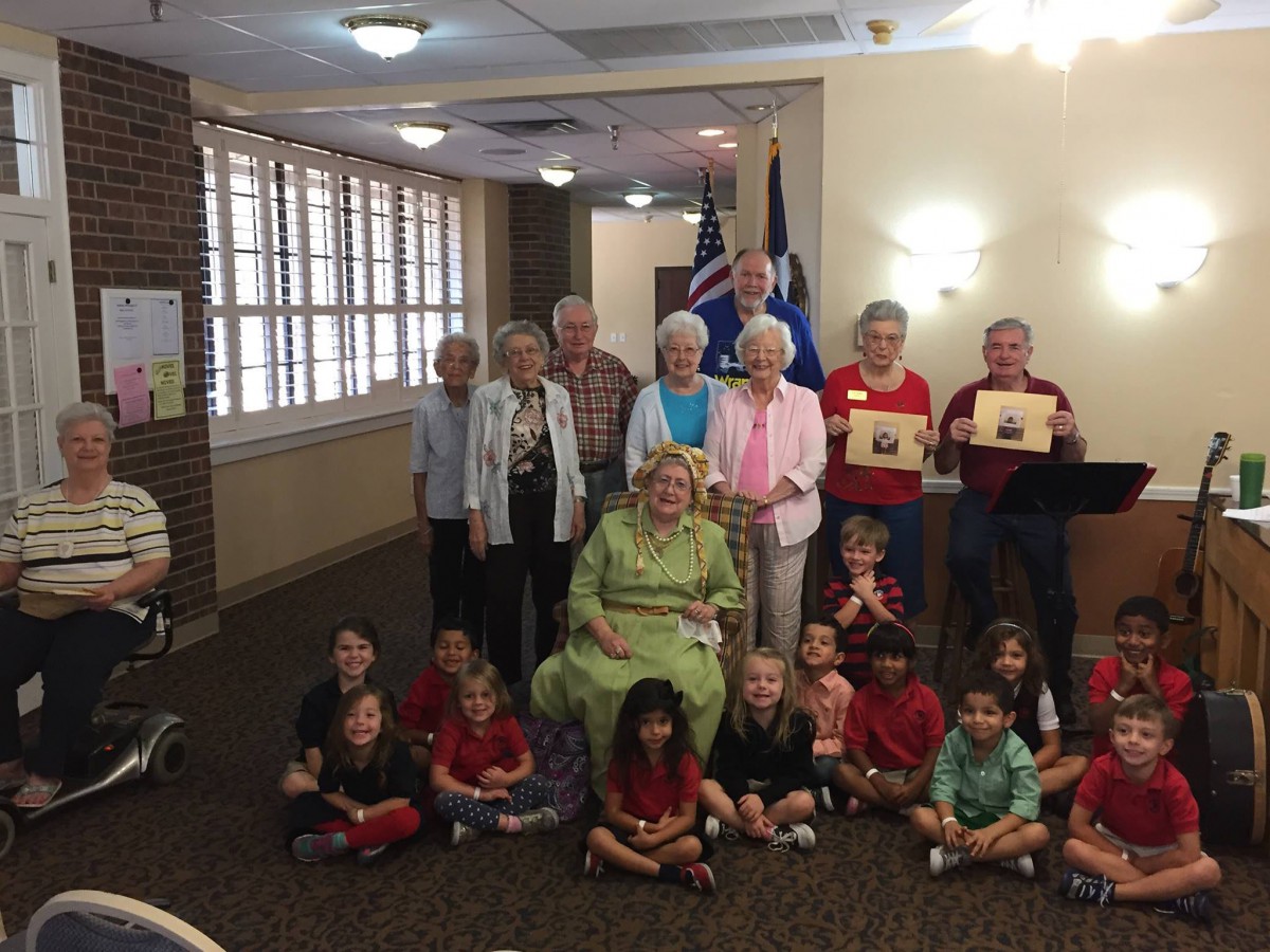 A group of children and senior citizens pose together indoors, with flags in the background, likely during a school event.