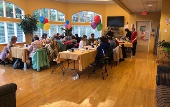 A group of people enjoying a festive gathering with balloons and dining tables in a bright indoor setting.