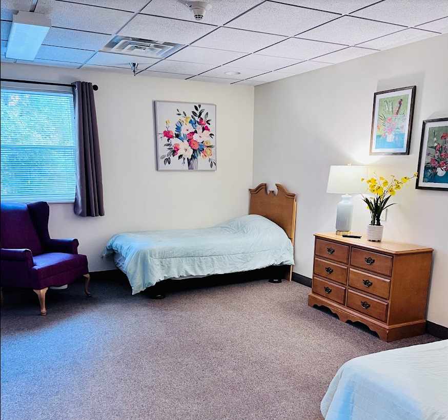 A well-lit bedroom with two single beds, a dresser with a lamp and flowers, and a comfortable chair near a window.