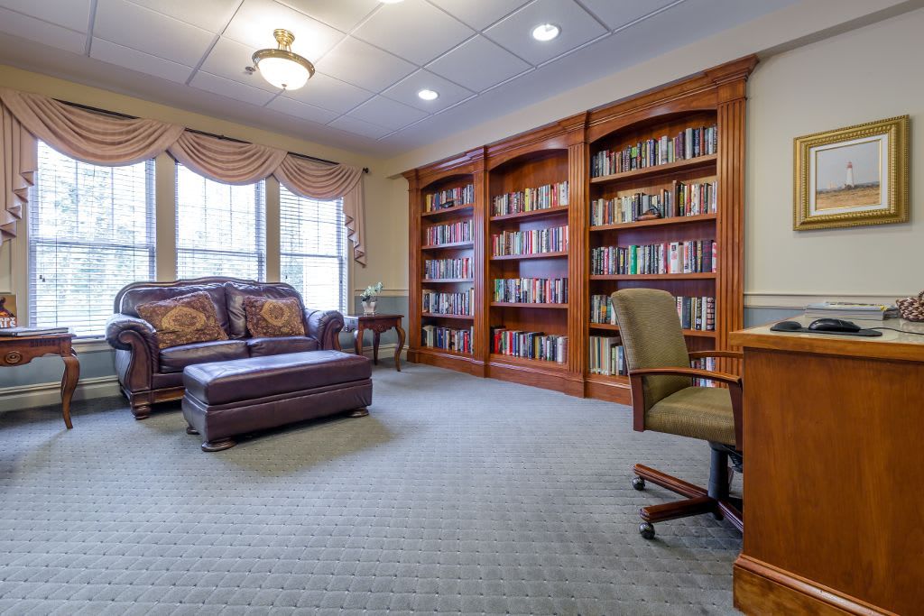A cozy library room with bookshelves, an armchair, ottoman, and a desk under soft lighting.