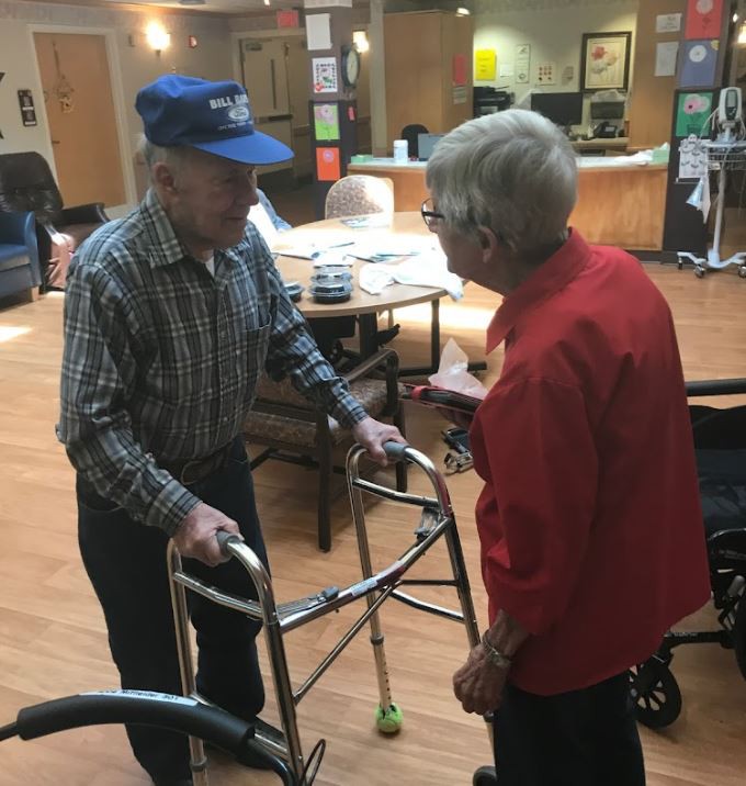 Two elderly individuals conversing indoors, one using a walker and the other in a wheelchair.