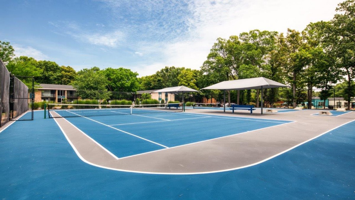 An outdoor tennis court with a blue surface, surrounded by trees and shaded seating areas.