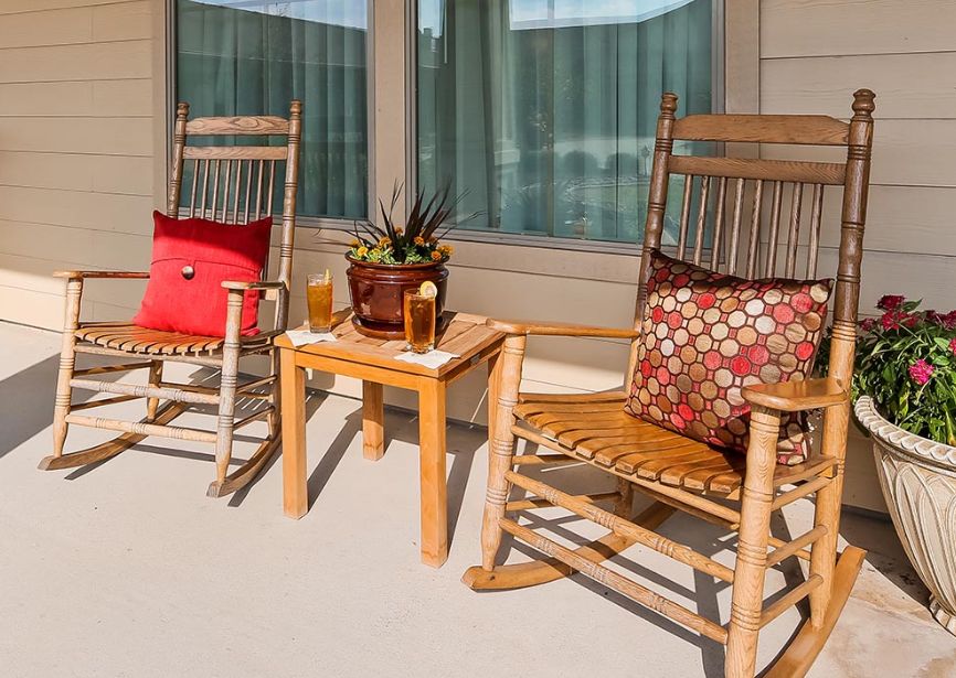 A cozy patio setup with two wooden rocking chairs, colorful pillows, a small side table holding drinks, and a potted plant.