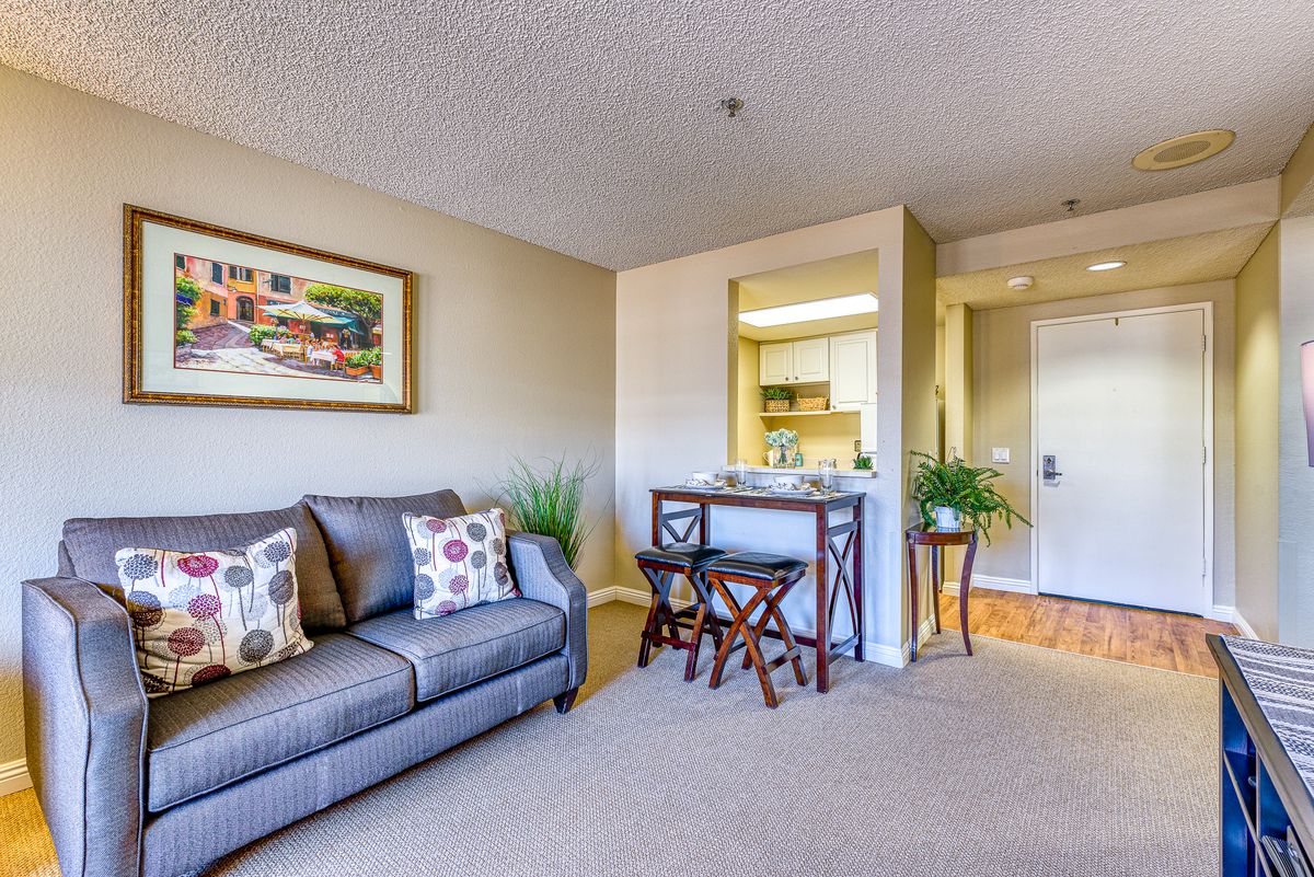 A cozy living room with a sofa, artwork, and plants, overlooking a kitchen area.