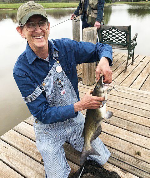 A smiling man in overalls holds a catfish on a dock.