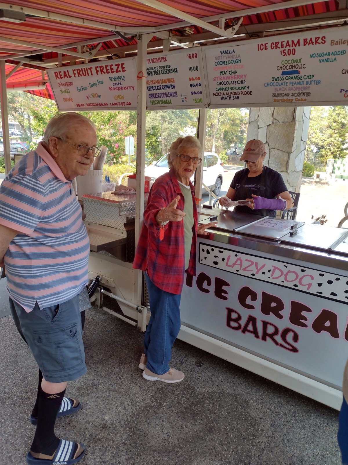 People enjoy a sunny day at an ice cream stand in the park.