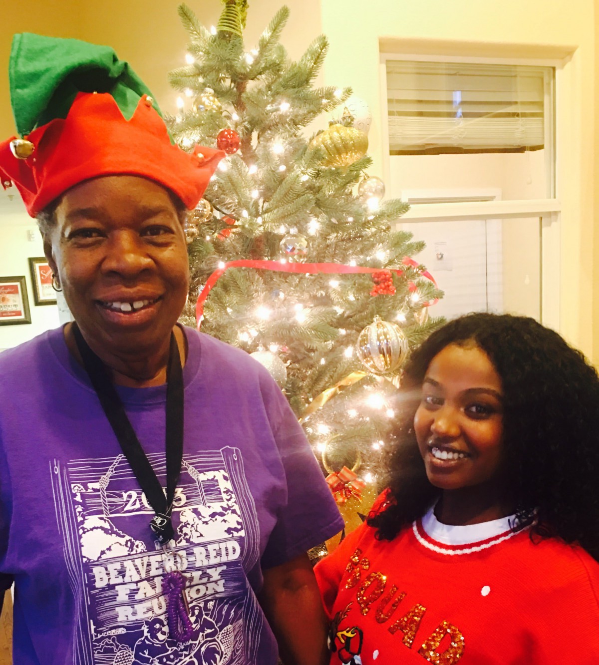 Two people smiling in front of a decorated Christmas tree, wearing festive hats.