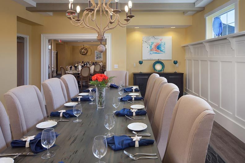 A beautifully set dining room with elegant decor, featuring a long table adorned with blue napkins and a centerpiece of red flowers under a grand chandelier.
