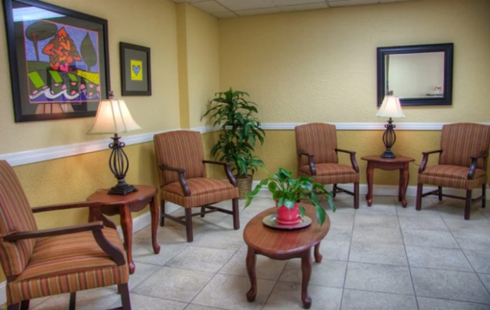 A cozy waiting room with comfortable chairs, a central table, lamps, and decorative plants.