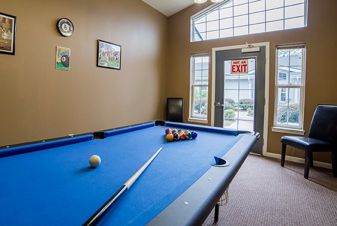 A game room featuring a pool table with blue felt and a cue stick, located near large windows and an exit door.