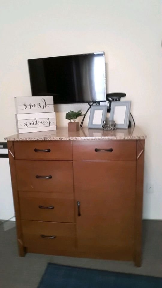 A wooden dresser topped with decorative items, including picture frames and a small plant, beneath a wall-mounted TV.