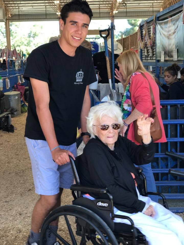 A young man is happily pushing an elderly woman in a wheelchair at an outdoor event.