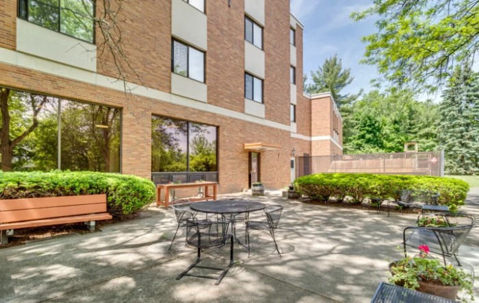 A sunny outdoor patio area with a table, chairs, and benches surrounded by a brick building and trees.