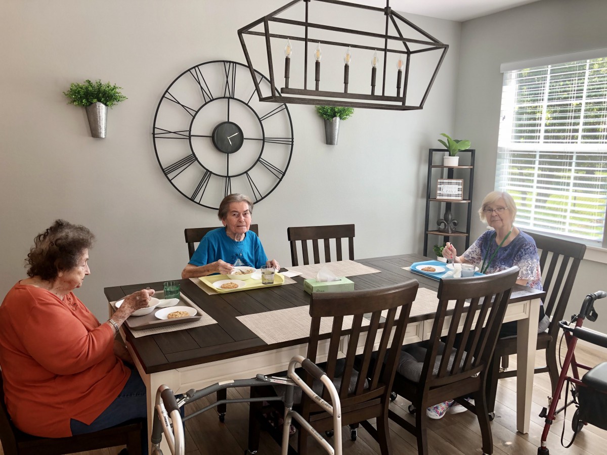 Three elderly individuals engage in an art activity around a dining table in a well-lit room.