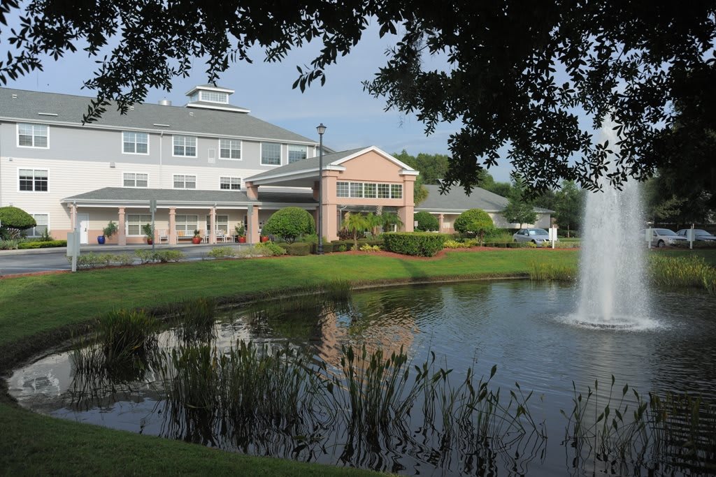 A scenic view of a building by a pond with a fountain and surrounding greenery.