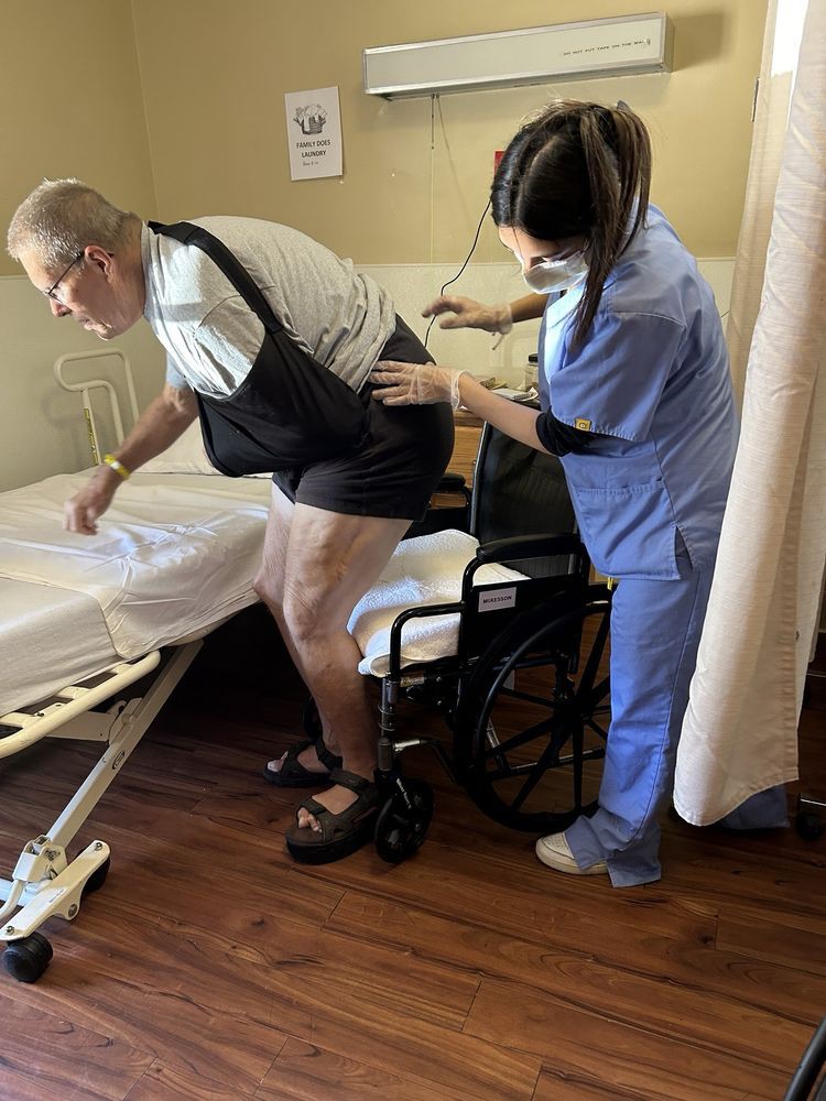 A nurse assists an elderly man transitioning from a wheelchair to a bed in a healthcare setting.