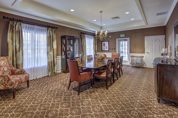A well-furnished dining room with a large table and upholstered chairs, featuring elegant interior design elements like a chandelier and patterned carpet.