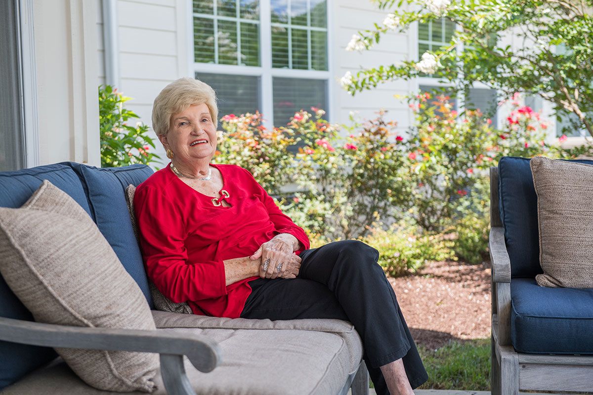 An elderly woman sitting comfortably on a patio chair outdoors, surrounded by a garden.