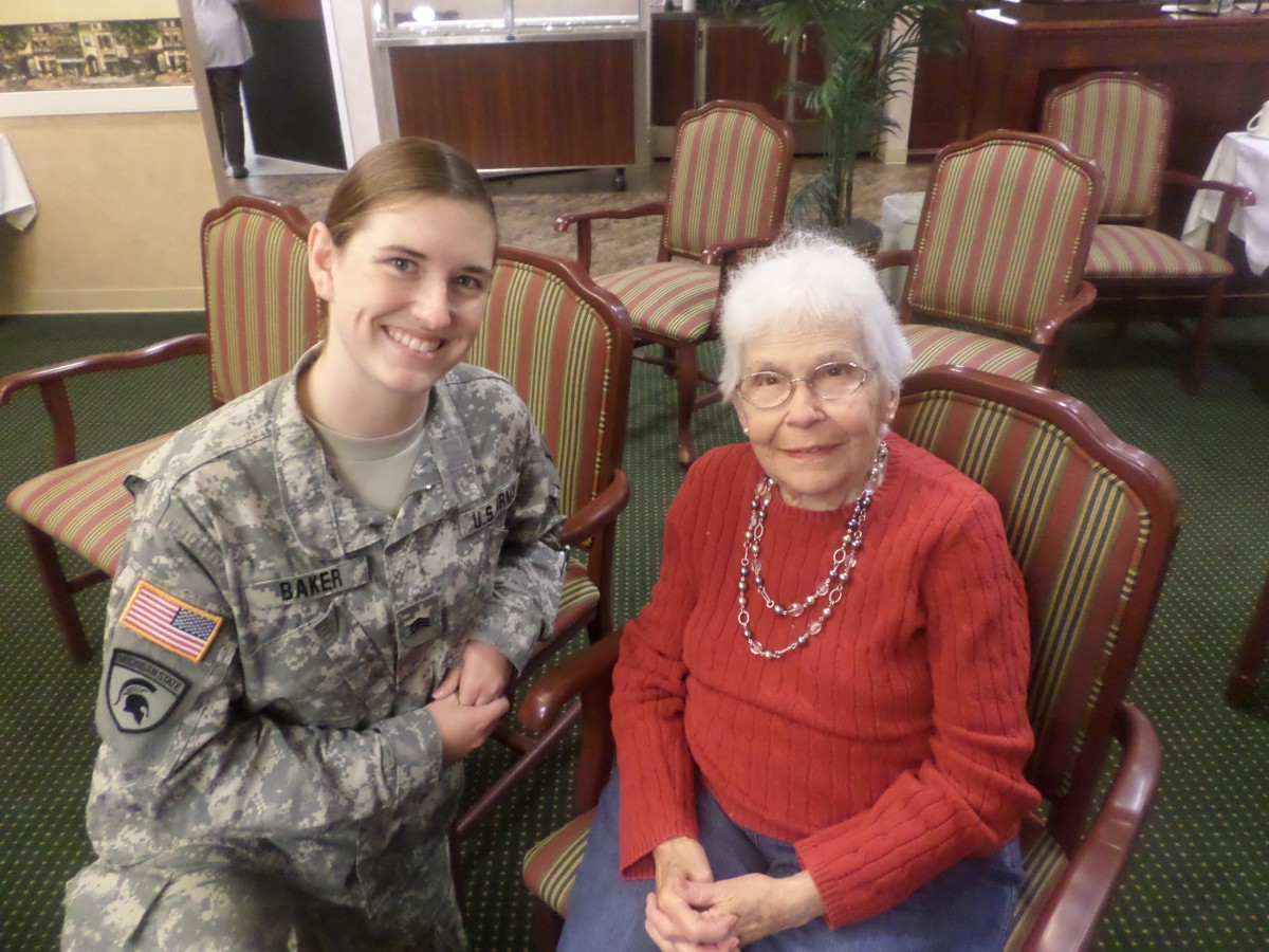 A soldier in uniform smiles while sitting next to an elderly woman indoors.