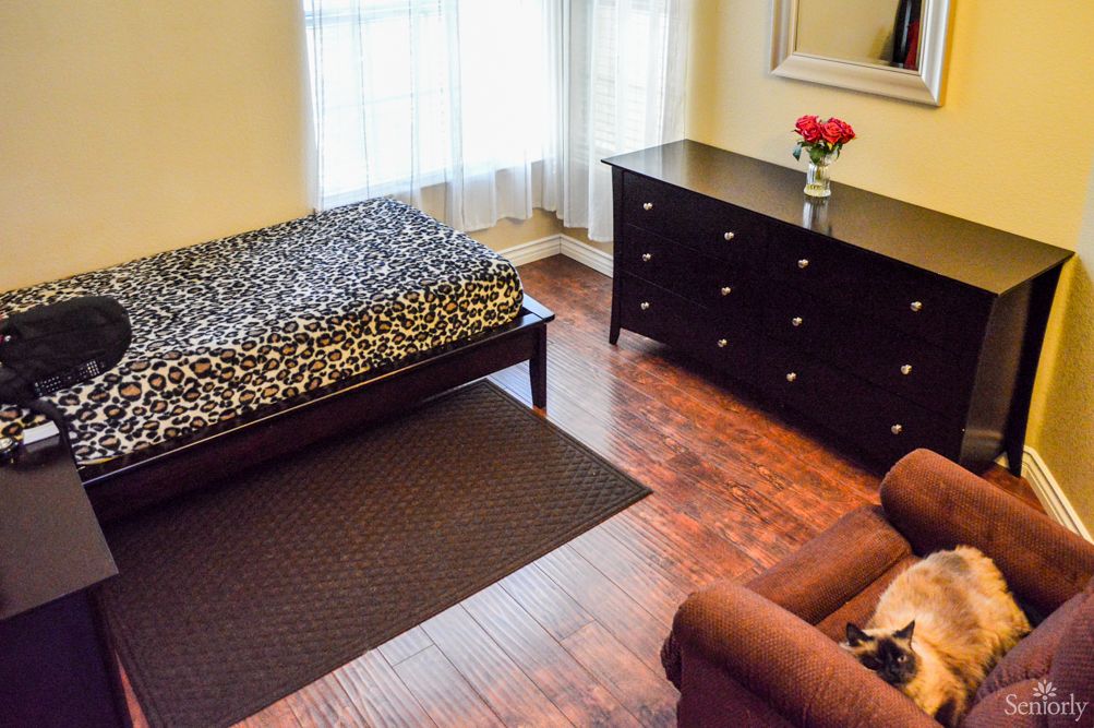 A cozy bedroom featuring a single bed with patterned bedding, a dark wooden dresser topped with a vase of roses, an armchair, and a large window letting in natural light.