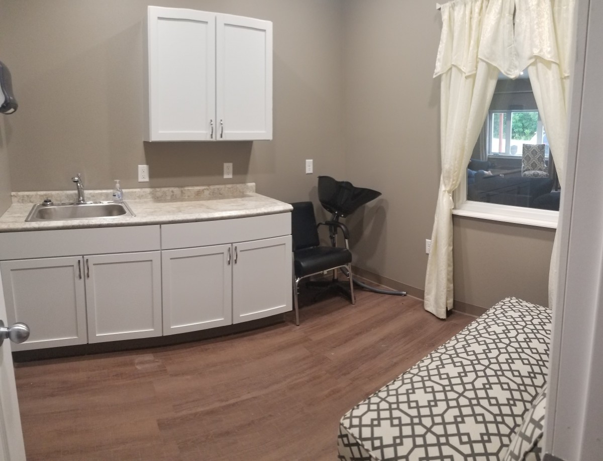 A small room featuring a sink with cabinets, a chair, a window with curtains, and part of a bed.