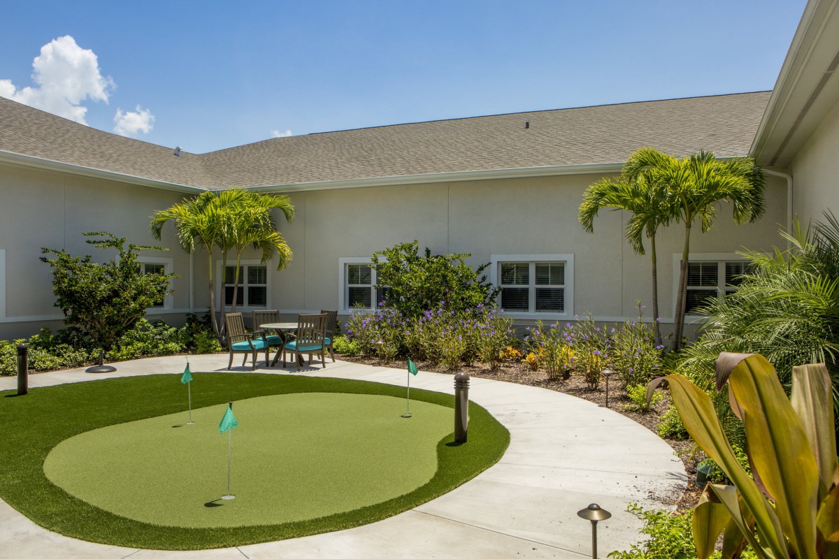 A putting green surrounded by lush landscaping and palm trees in a sunny outdoor patio area.